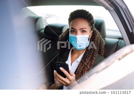 Woman in a sterile mask in taxi car on a back seat looking out of window checking her cell phone. Woman in a sterile mask in taxi car on a back seat looking out of window checking her cell phone. 101113817
