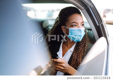 Woman in a sterile mask in taxi car on a back seat looking out of window checking her cell phone. 101113818