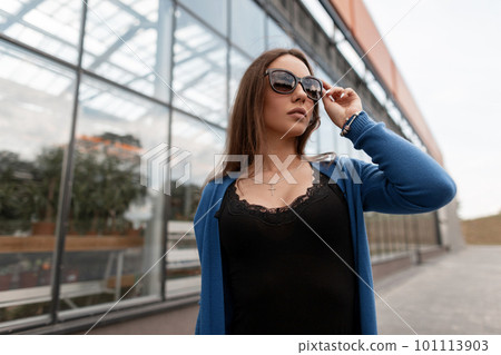Urban young pretty woman hipster in a knitted blue cape in vintage sunglasses in a trendy black t-shirt is standing near the glass building outdoors. Beautiful girl model in the city. Summer fashion. 101113903