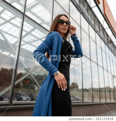 Glamorous hipster young woman in a vintage long knitted cape in jeans in a t-shirt in fashionable sunglasses walks on the street near the glass building. Attractive pretty stylish girl on the street. Glamorous hipster young woman in a vintage long knitted cape in jeans in a t-shirt in fashionable sunglasses walks on the street near the glass building. Attractive pretty stylish girl on the street. 101113918
