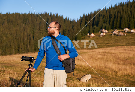 Happy man doing photo shoot in mountains. Professional photographer taking photos of hills at sunny day. Man holding his camera and bag on background of mountain forest. 101113933