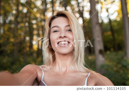 Happy young blond woman with a positive smile in a trendy pink dress makes a selfie on the background of a trees and a sunset in the forest. Pretty joyful girl resting in nature on a summer day. Happy young blond woman with a positive smile in a trendy pink dress makes a selfie on the background of a trees and a sunset in the forest. Pretty joyful girl resting in nature on a summer day. 101114105