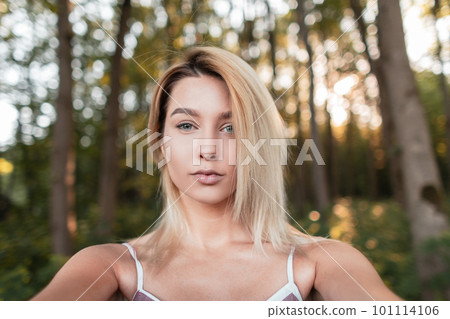 Beautiful stylish young woman blonde in a vintage pink striped dress resting in the woods on a sunny day and taking pictures of herself on the phone. Attractive girl makes selfie. 101114106