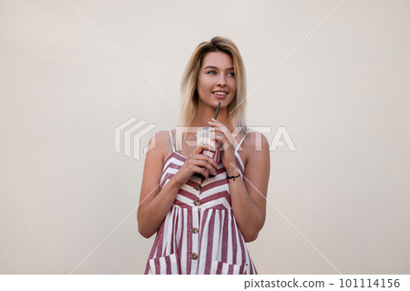 Happy pretty beautiful young blond woman in a stylish summer pink striped dress with a sweet cold milk drink rest near a vintage wall in the city. Joyful cute girl model enjoys outdoors travels. Happy pretty beautiful young blond woman in a stylish summer pink striped dress with a sweet cold milk drink rest near a vintage wall in the city. Joyful cute girl model enjoys outdoors travels. 101114156