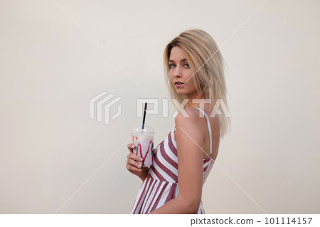 Glamorous beautiful young blond woman in a vintage pink striped dress with a sweet cold milk drink posing near a building in the city. Urban cute girl model enjoys outdoors walk. Good summer day. 101114157