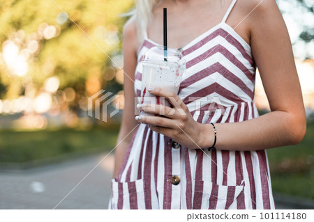 Close-up of a young woman in a summer pink striped dress with a milkshake in a sunny day outdoors in the park. Stylish girl enjoys a walk and a sweet drink outdoors in the park. 101114180