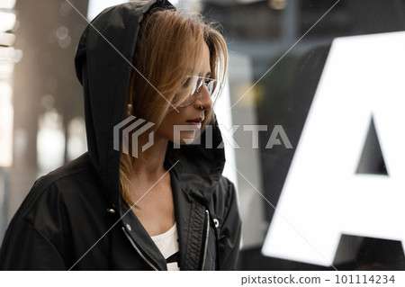 Sexy red-haired young woman hipster in stylish glasses in a fashionable hooded jacket is standing in a shopping center near a bright modern glass showcase. Beautiful girl on vacation. Youth style. Sexy red-haired young woman hipster in stylish glasses in a fashionable hooded jacket is standing in a shopping center near a bright modern glass showcase. Beautiful girl on vacation. Youth style. 101114234