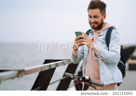 Young man with phone riding electric scooter on a pier near sea. Ecological transportation concept. Young man with phone riding electric scooter on a pier near sea. Ecological transportation concept. 101115005