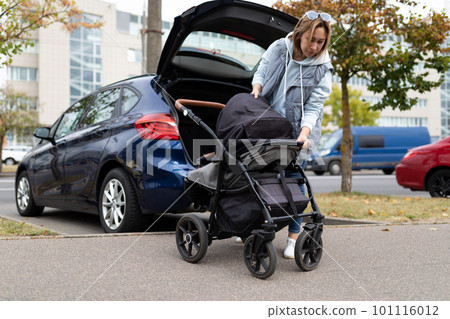 young woman parent folds the baby stroller into the luggage compartment of the car 101116012
