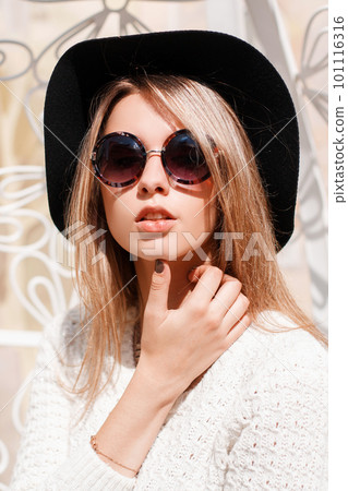 Portrait of an attractive young hipster woman in an elegant black hat in a vintage knitted white sweater in round sunglasses with a necklace on a white background. Fashionable European girl. Portrait of an attractive young hipster woman in an elegant black hat in a vintage knitted white sweater in round sunglasses with a necklace on a white background. Fashionable European girl. 101116316