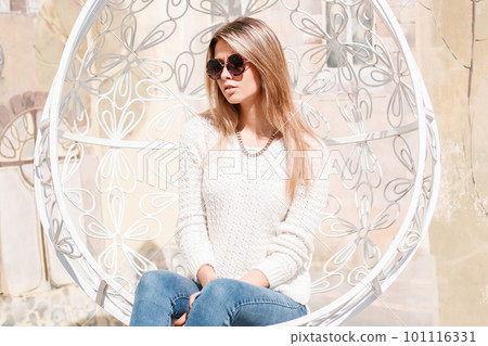 European stylish young woman hipster in sunglasses with a brilliant necklace in stylish jeans in a knitted sweater sits in a vintage metal white hanging chair. Happy girl enjoys the sun. European stylish young woman hipster in sunglasses with a brilliant necklace in stylish jeans in a knitted sweater sits in a vintage metal white hanging chair. Happy girl enjoys the sun. 101116331