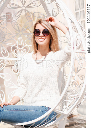 Happy cute young hipster woman in a knitted sweater in stylish jeans with black round sunglasses sits and smiles in a hanging beautiful metal chair. Charming cheerful girl resting. 101116337