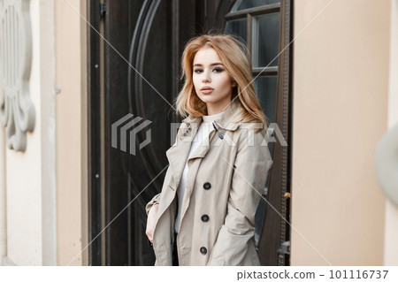 Beautiful stylish young blond woman in an autumn coat in a white t-shirt with a black leather stylish bag stands against the background of an old brown wooden hotel door. Fashionable american girl. Beautiful stylish young blond woman in an autumn coat in a white t-shirt with a black leather stylish bag stands against the background of an old brown wooden hotel door. Fashionable american girl. 101116737