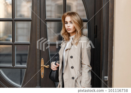 Stylish attractive young blond woman in an autumn coat with a black leather stylish bag stands near a vintage wooden mirror door. Cute girl Stylish attractive young blond woman in an autumn coat with a black leather stylish bag stands near a vintage wooden mirror door. Cute girl 101116738