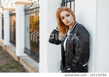 Beautiful young blonde woman in a black summer stylish jacket in black jeans in a white polo shirt standing near a vintage metal fence. Fashionable modern girl. 101116769
