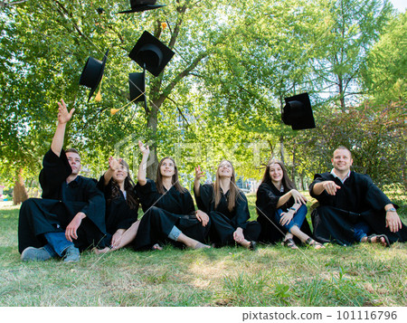 Happy student friends in mantle sit on the grass in the university park. Graduates throw their academic cap up Happy student friends in mantle sit on the grass in the university park. Graduates throw their academic cap up 101116796
