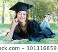 Young smiling female student in black graduate gown lies on the grass in the university park 101116859