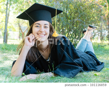 Young smiling female student in black graduate gown lies on the grass in the university park 101116859