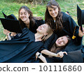 Several young smiling female students in black graduation gowns lie on the grass in the university park 101116860