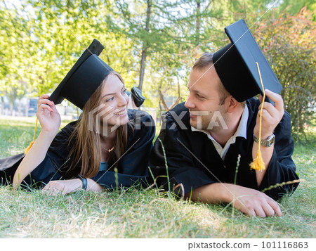 Happy guy and girl look at each other lying on the lawn in a black graduate gown and an academic cap of the university 101116863