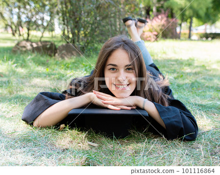 Portrait of a happy smiling girl in a black robe lying on the grass. A young woman is celebrating her graduation. New life 101116864