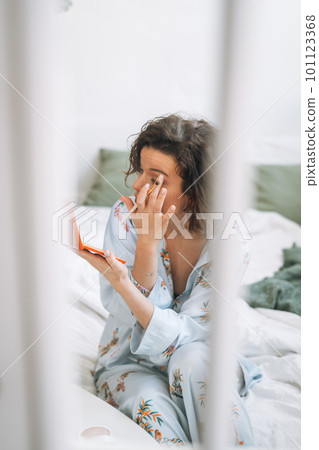 Young brunette woman in blue pajamas applies makeup with eye shadow palette in hands at home Young brunette woman in blue pajamas applies makeup with eye shadow palette in hands at home 101123368