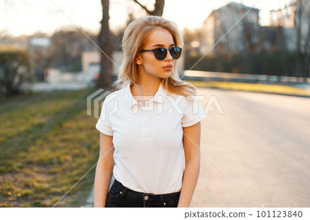 Young attractive hipster woman in stylish sunglasses in a white polo t-shirt in black jeans walking in downtown at sunset. Cute stylish girl. Summer evening. Young attractive hipster woman in stylish sunglasses in a white polo t-shirt in black jeans walking in downtown at sunset. Cute stylish girl. Summer evening. 101123840