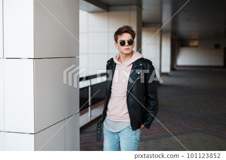 Fashionable young hipster man in black sunglasses in stylish black jacket in pink sweatshirt in trendy jeans in a gray t-shirt is standing in the indoors. Modern guy with a stylish hairstyle. Fashionable young hipster man in black sunglasses in stylish black jacket in pink sweatshirt in trendy jeans in a gray t-shirt is standing in the indoors. Modern guy with a stylish hairstyle. 101123852
