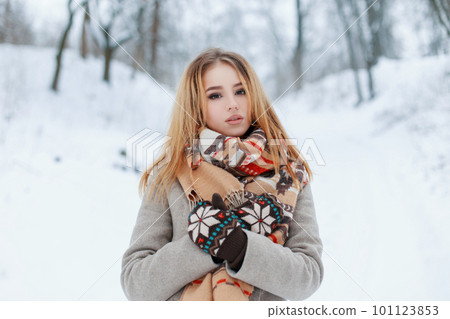 Beautiful attractive young woman in a fashionable gray coat in knitted vintage mittens with a stylish beige scarf with a pattern rest in a snowy park on a winter cold day. Charming girl blonde. Beautiful attractive young woman in a fashionable gray coat in knitted vintage mittens with a stylish beige scarf with a pattern rest in a snowy park on a winter cold day. Charming girl blonde. 101123853
