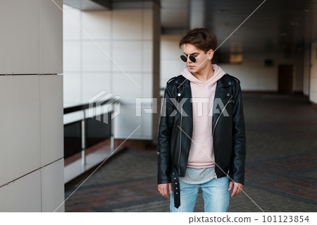 Fashionable young hipster man in black sunglasses in stylish black jacket in pink sweatshirt in trendy jeans in a gray t-shirt is standing in the room. Serious cool guy with a beautiful hairstyle Fashionable young hipster man in black sunglasses in stylish black jacket in pink sweatshirt in trendy jeans in a gray t-shirt is standing in the room. Serious cool guy with a beautiful hairstyle 101123854