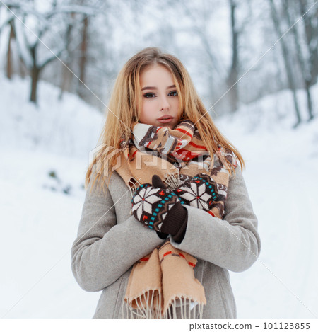 Modern charming young girl in a vintage warm coat in beautiful woolen mittens with a fashionable scarf with a red pattern posing outdoors on the background of snowy trees in the park. Stylish girl. Modern charming young girl in a vintage warm coat in beautiful woolen mittens with a fashionable scarf with a red pattern posing outdoors on the background of snowy trees in the park. Stylish girl. 101123855