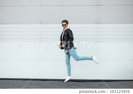 Attractive young hipster man in black glasses in a fashionable leather black jacket in a light pink sweatshirt in blue jeans in white sneakers shows off the jump near the white wall. Cute american guy Attractive young hipster man in black glasses in a fashionable leather black jacket in a light pink sweatshirt in blue jeans in white sneakers shows off the jump near the white wall. Cute american guy 101123856