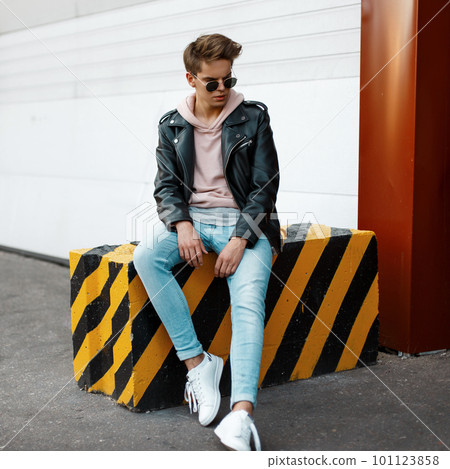 Stylish handsome young man in black sunglasses in fashionable clothes with a hairstyle sitting on a striped black-yellow concrete slab in the outdoors near the white metal gates. American modern guy Stylish handsome young man in black sunglasses in fashionable clothes with a hairstyle sitting on a striped black-yellow concrete slab in the outdoors near the white metal gates. American modern guy 101123858