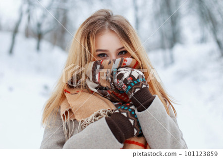 Young happy woman with beautiful brown eyes in winter fashionable warm clothes in vintage mittens in the winter forest. Funny stylish girl with a scarf on her face. 101123859