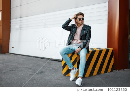 Young hipster man in a pink sweatshirt in trendy sunglasses in a black jacket in jeans in white sneakers is sitting on a striped concrete slab against the background of a metal white gate.American guy Young hipster man in a pink sweatshirt in trendy sunglasses in a black jacket in jeans in white sneakers is sitting on a striped concrete slab against the background of a metal white gate.American guy 101123860