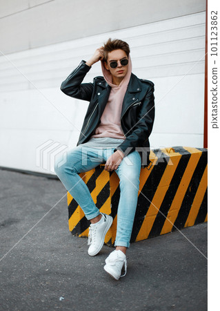 Stylish American young hipster man in a pink sweatshirt in trendy sunglasses in a leather jacket in jeans in white sneakers sitting on a striped concrete slab against a white gate. Cool guy Stylish American young hipster man in a pink sweatshirt in trendy sunglasses in a leather jacket in jeans in white sneakers sitting on a striped concrete slab against a white gate. Cool guy 101123862