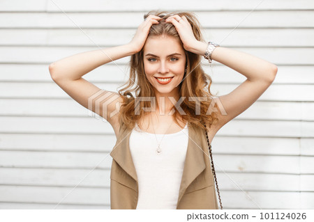 Cheerful young woman in an elegant summer beige vest in a stylish T-shirt posing near a wooden vintage white wall. Joyful girl with a beautiful smile. Positive mood 101124026