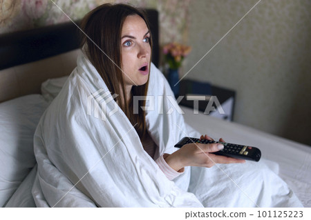 Young woman covered blanket relaxing in bed in the evening and watching tv Young woman covered blanket relaxing in bed in the evening and watching tv 101125223