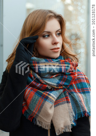 Cute charming young woman with blond hair in a stylish winter coat with a woolen vintage scarf in black gloves posing on the background of bright festive garlands. Sweet girl waiting for the magic 101128178