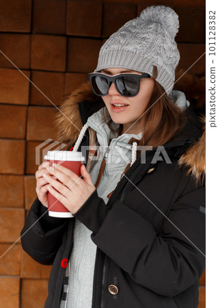 Stylish european young hipster woman in a knitted hat in a warm sweatshirt in a jacket with fur in sunglasses near a vintage wooden wall holding a red cup with hot drink. Beautiful modern girl. 101128382