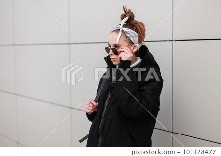 Young woman hipster in a stylish long jacket with a leather backpack with a stylish hairstyle in a trendy bandana in black sunglasses near a white wall in the outdoors. American girl. Modern fashion. 101128477