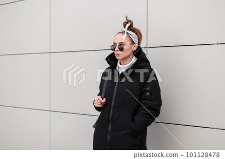 Modern young hipster woman in round sunglasses in a stylish coat in a fashionable bandana with a leather backpack is standing near a white wall a warm winter day. Fashionable american girl on vacation 101128478