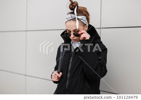 Modern young hipster woman with stylish hairstyle in a bandana in a fashionable black jacket with a stylish backpack in a knitted golf in stylish sunglasses posing near a white building. Fashion girl. Modern young hipster woman with stylish hairstyle in a bandana in a fashionable black jacket with a stylish backpack in a knitted golf in stylish sunglasses posing near a white building. Fashion girl. 101128479