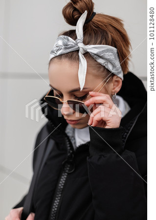 Young hipster woman in trendy sunglasses in a stylish bandana in a winter warm coat posing in a city near a white wall. American fashion girl. Young hipster woman in trendy sunglasses in a stylish bandana in a winter warm coat posing in a city near a white wall. American fashion girl. 101128480