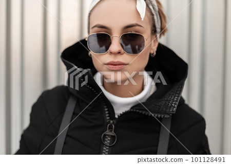 Portrait of a fashionable young hipster woman in trendy sunglasses with a bandana in white golf in a stylish jacket with a backpack on the background of a metal wall. Modern girl. 101128491