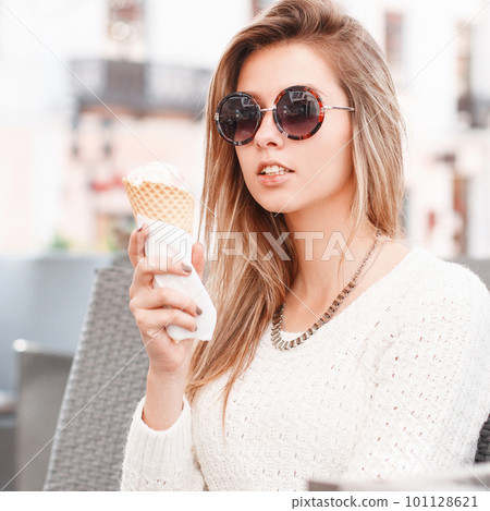 Charming young hipster blond woman in stylish sunglasses in a vintage knitted sweater with sweet ice cream sits in a cafe on a warm spring day. Cute girl eating a delicious ice cream. Charming young hipster blond woman in stylish sunglasses in a vintage knitted sweater with sweet ice cream sits in a cafe on a warm spring day. Cute girl eating a delicious ice cream. 101128621