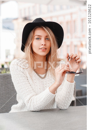 Cute attractive young woman in a white vintage knitted sweater in an elegant hat sitting on a warm spring day in an outdoor cafe. Charming joyful girl blonde. Cute attractive young woman in a white vintage knitted sweater in an elegant hat sitting on a warm spring day in an outdoor cafe. Charming joyful girl blonde. 101128622