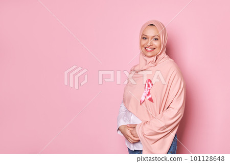 Smiling Middle-Eastern Muslim pregnant woman in pink hijab, wearing satin ribbon, emblem of fighting breast cancer and showing support for cancer survivors and patients, isolated over pink background 101128648