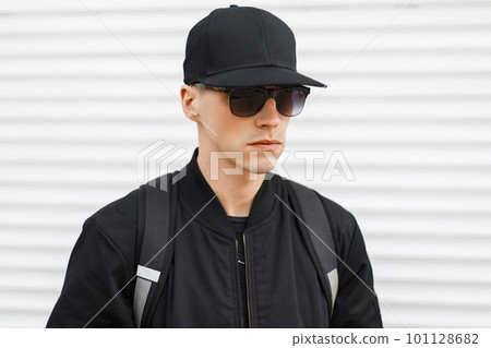 Stylish attractive hipster man in a black basketball cap in a fashionable black sunglasses in a trendy jacket with a black backpack on his shoulders near the metal white wall. American guy. 101128682