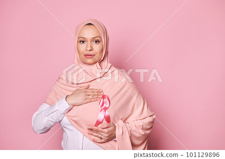 Confident Arab Muslim woman in hijab, wearing pink satin awareness ribbon, emblem of fighting Breast Cancer and supporting patients and survivors, looking confidently at camera on isolated background 101129896
